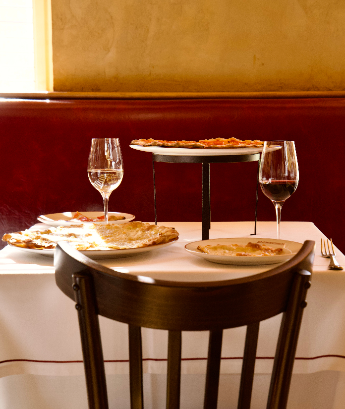 A white-tablecloth dining setup with flatbreads or pizza served on a raised stand and plates, paired with red and white wine in a warm booth setting.