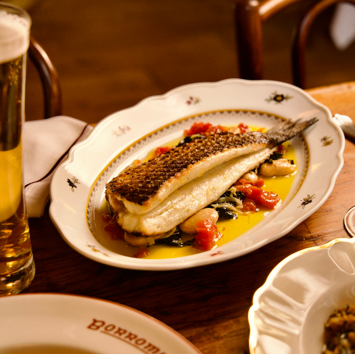 A crispy-skinned fish fillet on a vintage floral-rimmed oval plate with tomatoes, white beans, and a golden broth, served alongside a glass of beer.