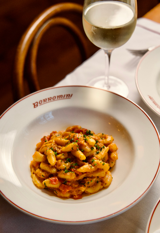 A bowl of crab pasta in a rich tomato-based sauce, garnished with herbs, served in a branded Borromini dish with a glass of white wine on a white tablecloth.