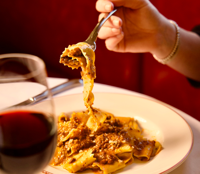 A person eats spaghetti with a fork over a white bowl, next to a glass of red wine on a small table.