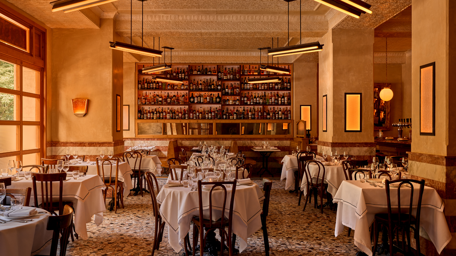 Elegant restaurant with wine bottle-lined walls, marble counter, and white tablecloths on wooden floor.