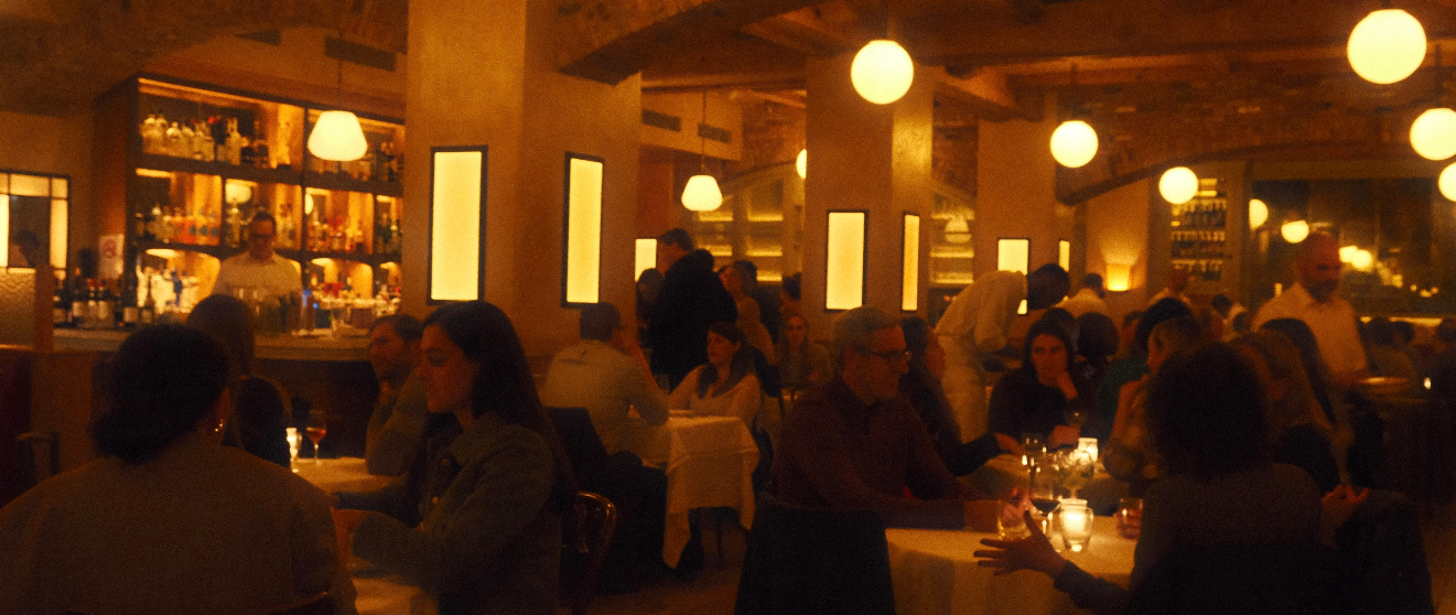 A warmly lit, bustling restaurant dining room with guests seated at white-tablecloth tables, glowing globe pendant lights overhead, and a backlit bar lined with bottles.
